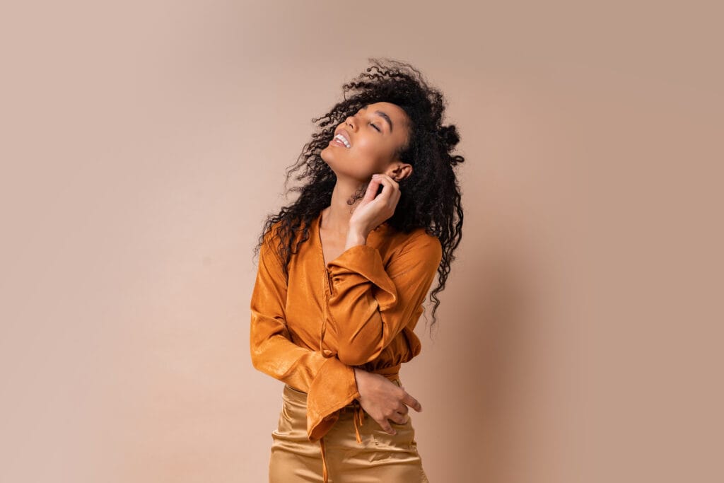 Glad african woman with perfect curly hairs in casual orange blouse and golden pants posing on beige background blog cover image